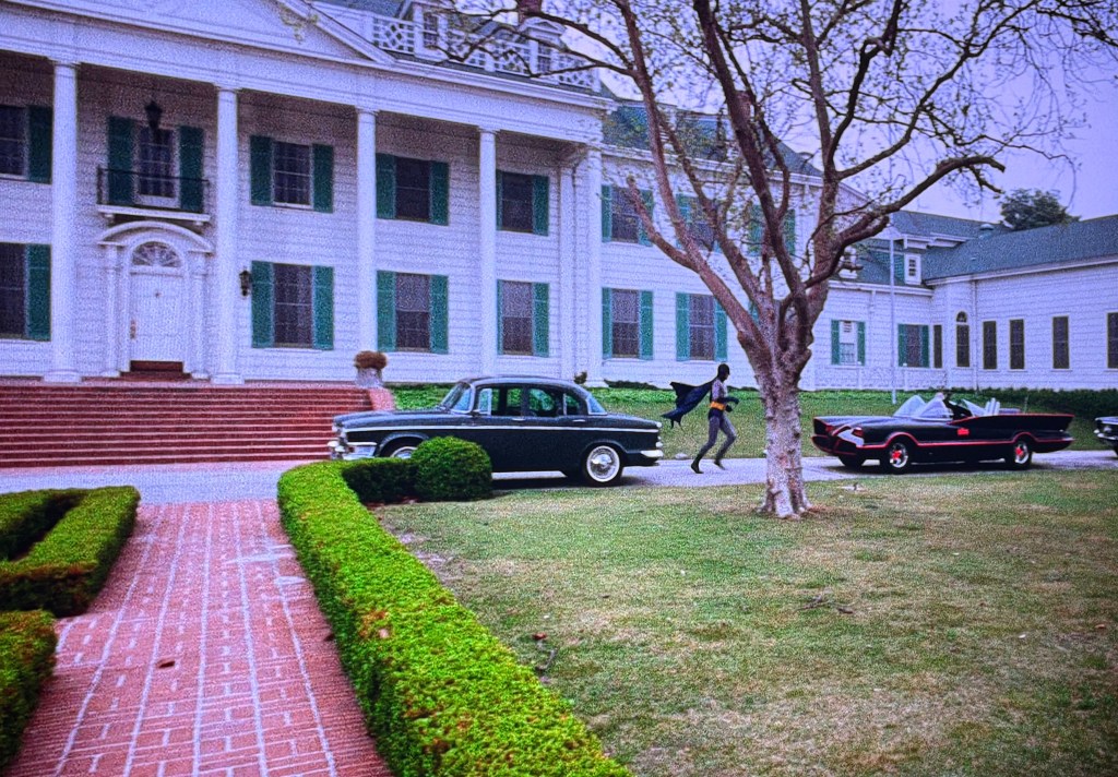 A large white mansion with green shutters, featuring a set of stairs at the entrance. In the foreground, a man in a dark outfit runs along a pathway lined with hedges, while two cars, a vintage black sedan and a red convertible, are parked nearby.