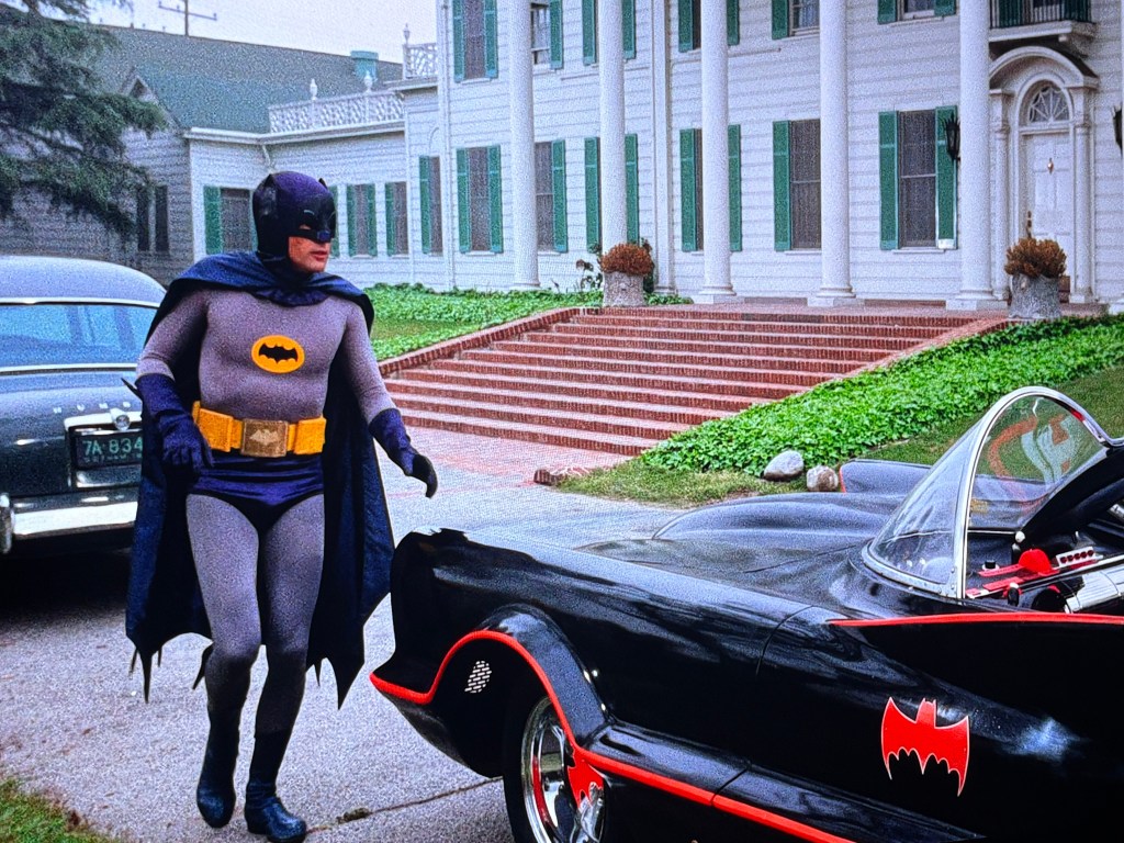 A man dressed as Batman stands beside a classic black Batmobile in front of a large mansion with green shutters and a well-kept garden.