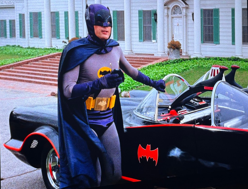 Batman in his iconic costume stands next to the Batmobile in front of a house with green shutters.