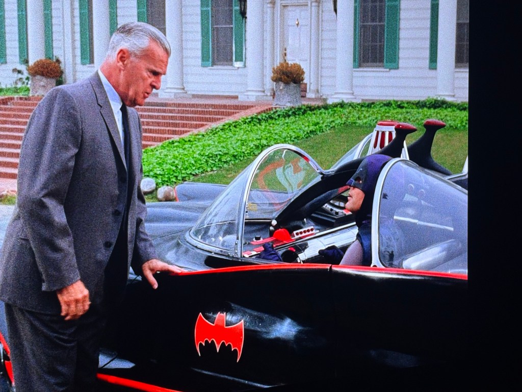 A scene featuring a man in a suit speaking to a character in a Batmobile, with a grand house and greenery in the background.