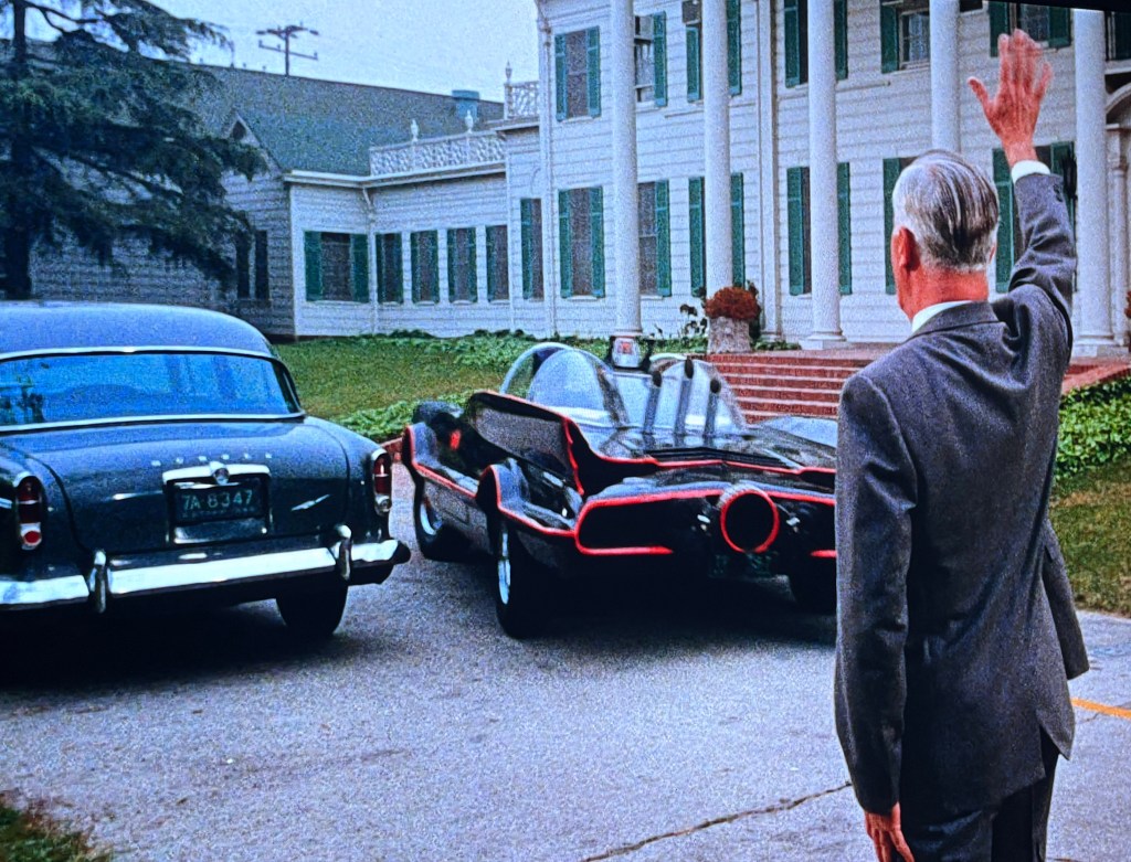 A man in a suit waves goodbye as a sleek black car with red accents, resembling a famous fictional vehicle, drives away in front of a large white house with green shutters.