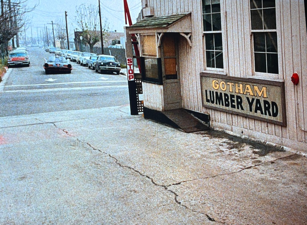 A street view of Gotham Lumber Yard featuring a wooden building with a prominent sign. In the foreground, there's a stop sign and a concrete sidewalk, while parked cars line the street in the background under a cloudy sky.