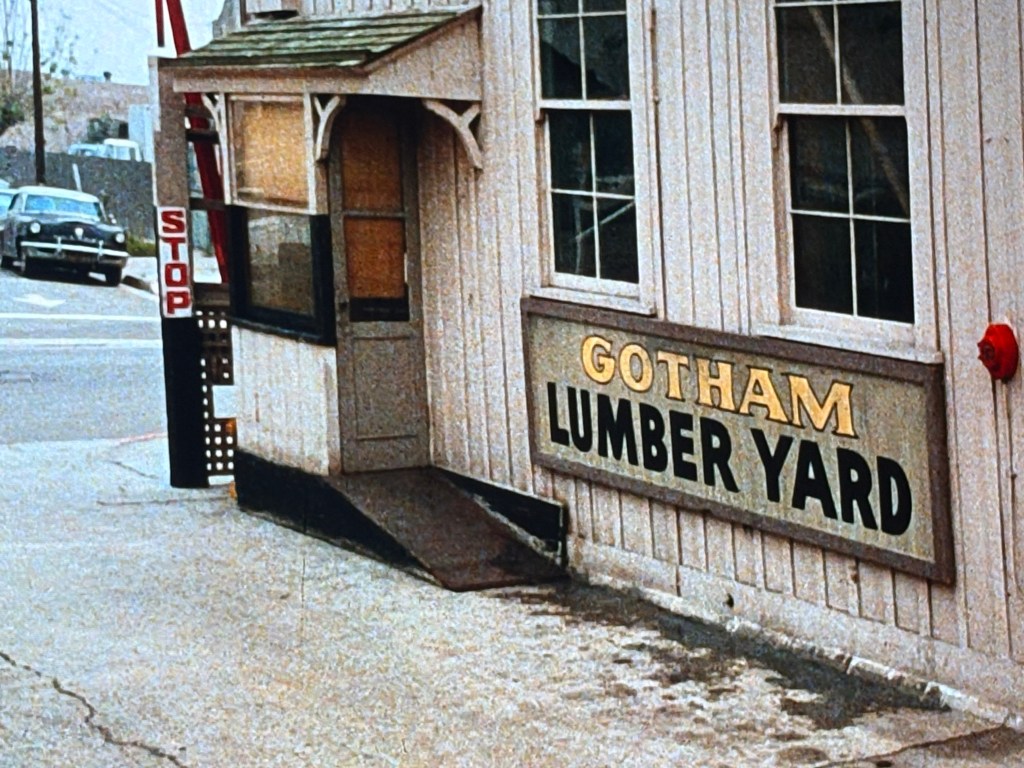 Exterior view of Gotham Lumber Yard with a wooden sign and a stop sign nearby.