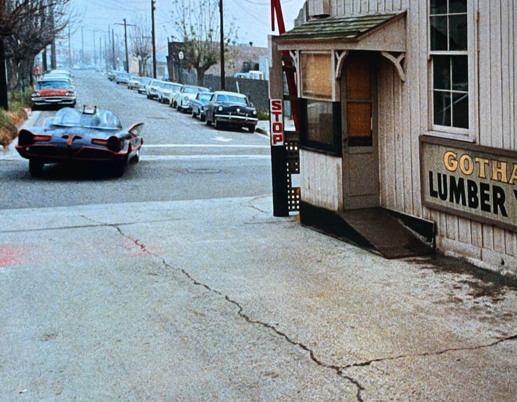 A black and red vehicle departs from a lumber yard labeled 'Gotham Lumber Warehouse,' on a cloudy street lined with parked cars.