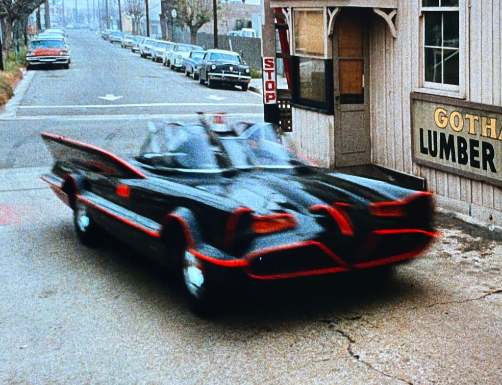 A black car with red accents resembling a superhero vehicle speeds past a lumber store, with parked cars lining the street.