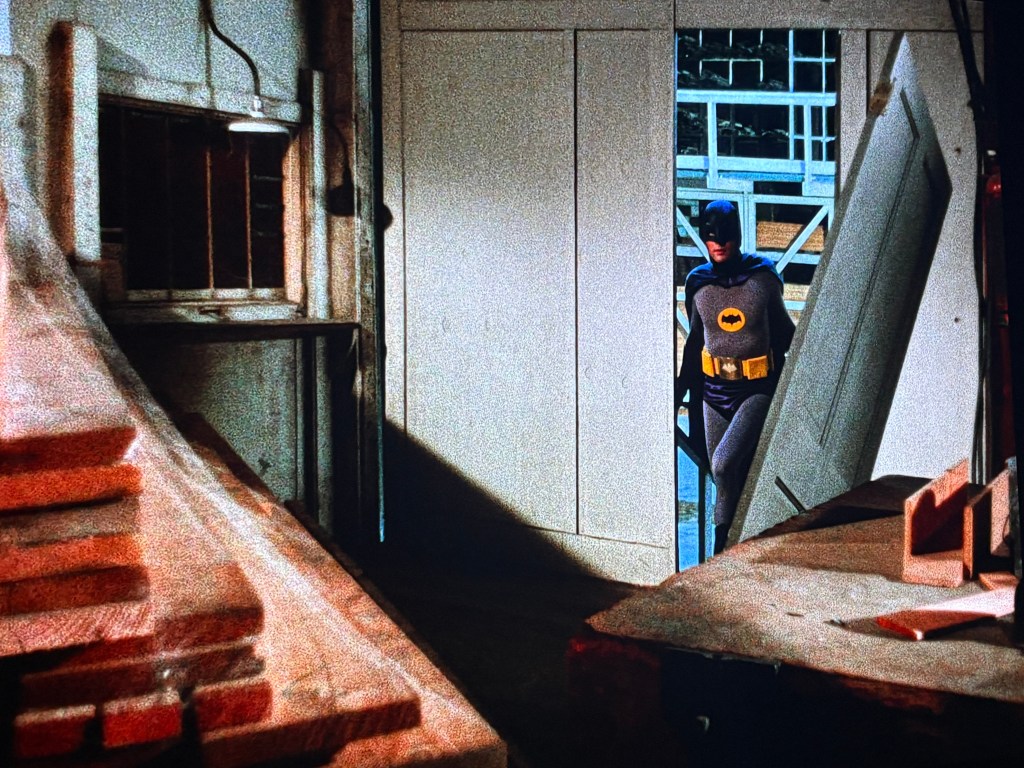A person dressed as Batman stands in a doorway, partially visible in a dimly lit room surrounded by wooden planks and construction materials.