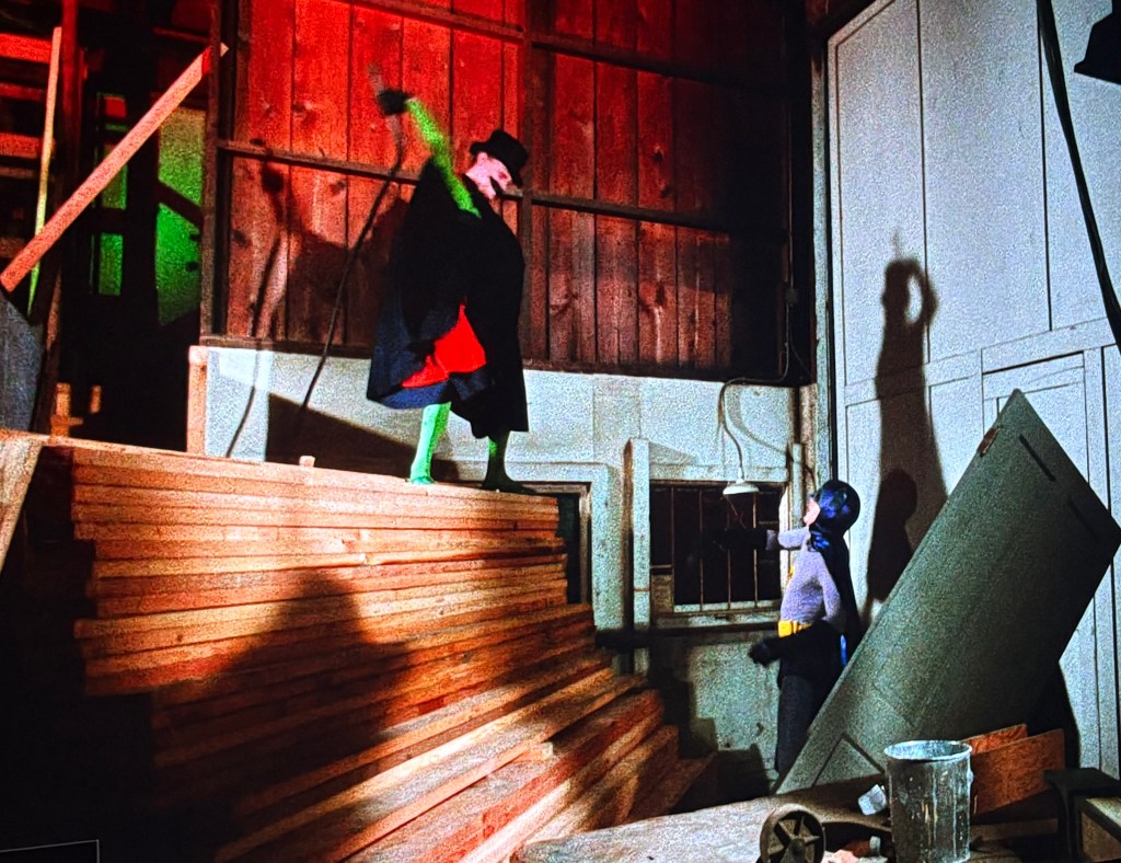 A performer in a green face paint and a black cape poses dramatically on wooden crates, while a person in a grey and black costume stands below looking up, with colorful lighting creating distinct shadows.