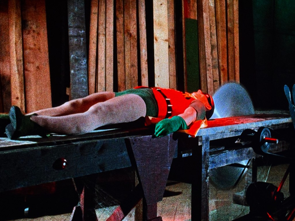 A character dressed in a red shirt, green shorts, and gloves, lies on a wooden table in a workshop-like setting, with saw blades visible in the background.