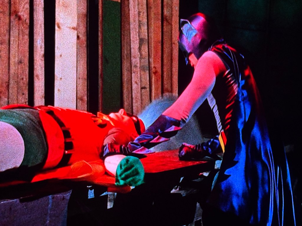 A scene featuring a superhero in a cape leaning over a figure dressed in a red and green costume lying on a wooden table, set against a backdrop of wooden planks.