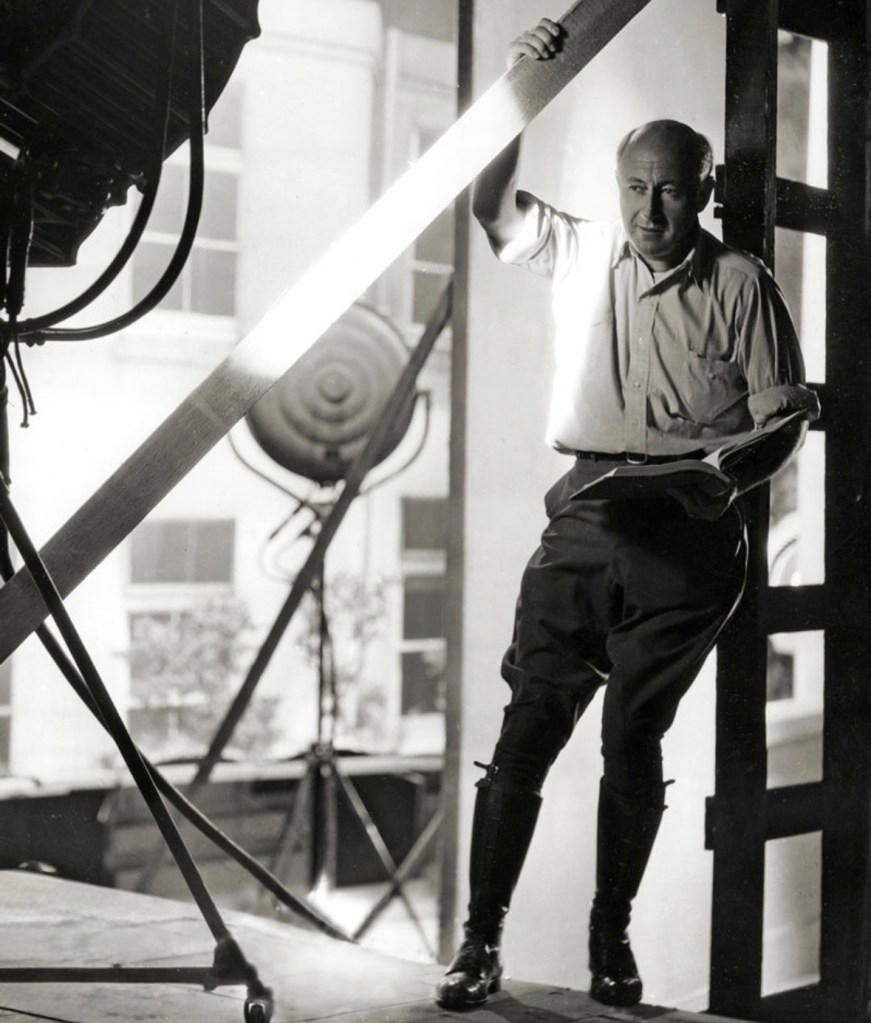 A man standing in a film studio, holding a script and leaning against a wooden beam, with studio lights and a backdrop visible.