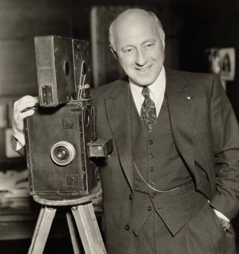 A smiling man in a suit stands next to a vintage camera on a tripod, exuding a sense of professionalism and nostalgia.