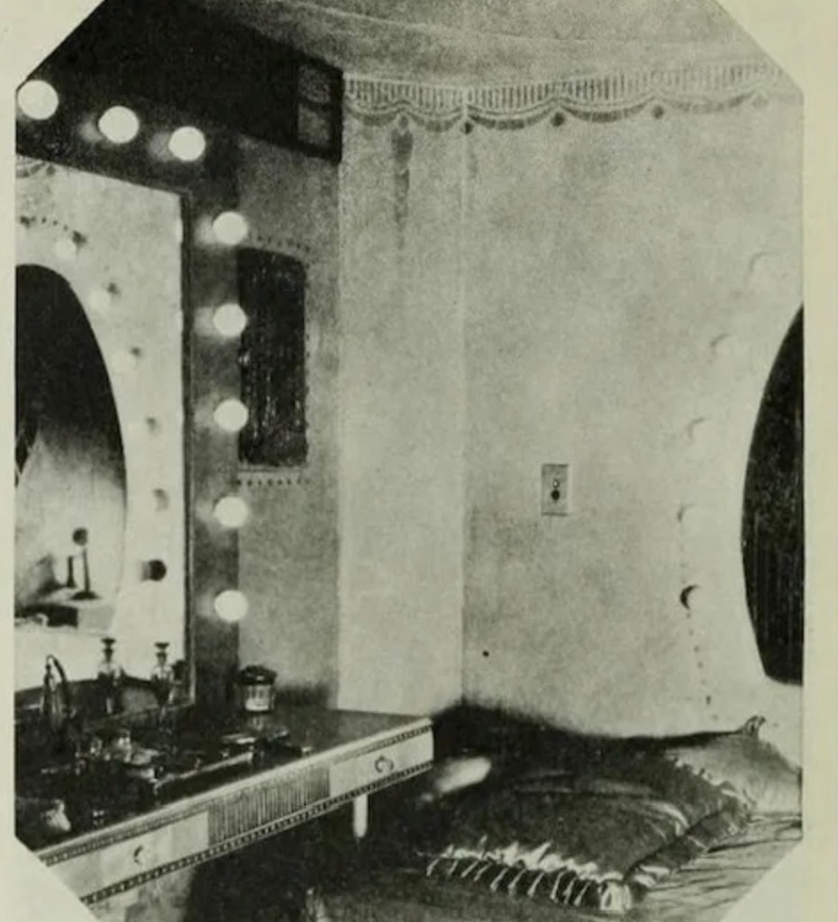 Vintage dressing room with a large mirror surrounded by lights, featuring a vanity table with various beauty products and a cushioned seating area.