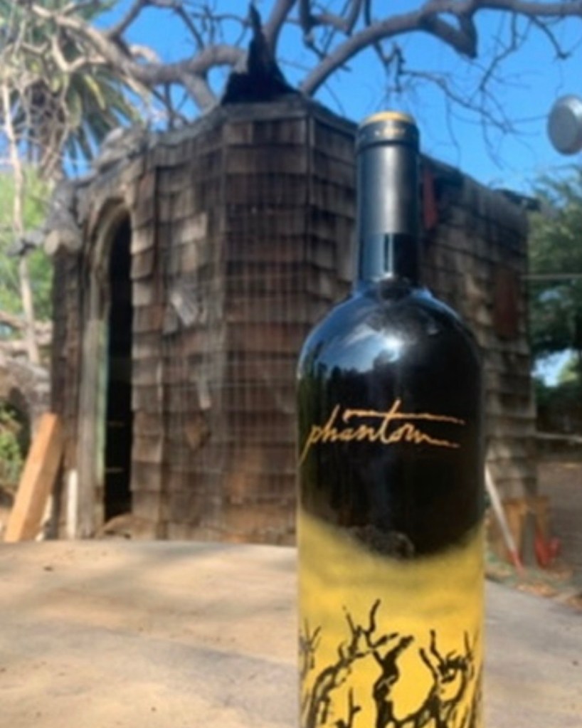A bottle of Phantom wine placed on a rustic surface with a wooden shed and trees in the background.