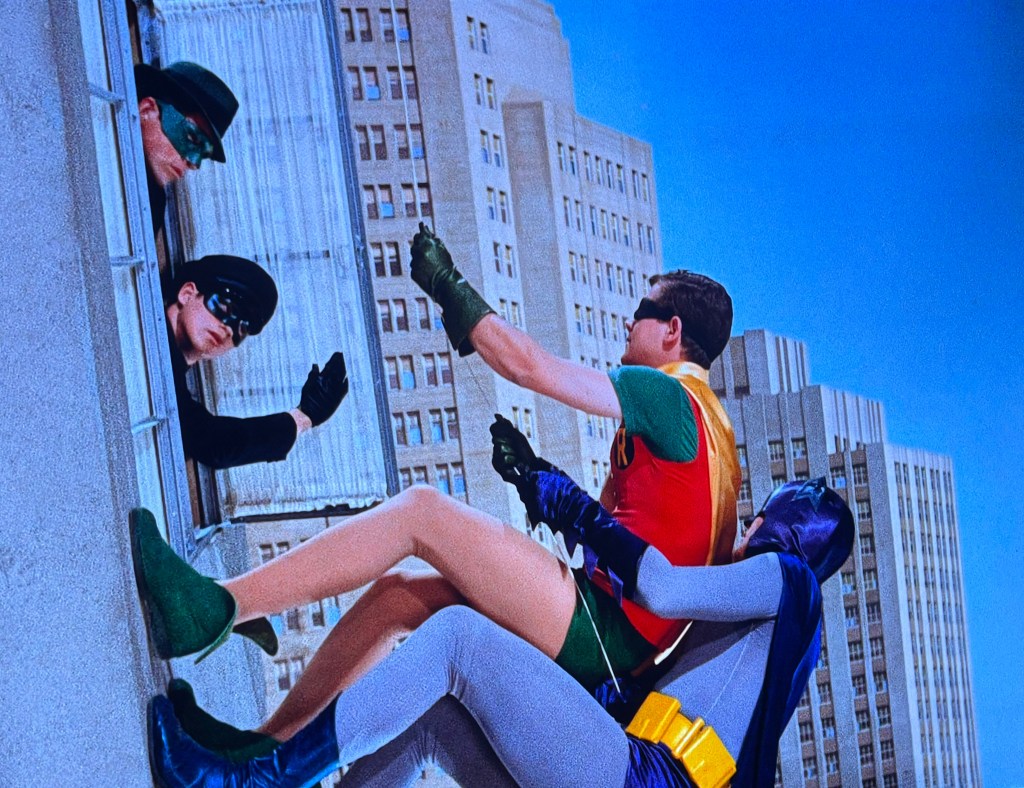A scene featuring a superhero duo performing a daring rescue on a building ledge, with a mysterious figure observing from a window.