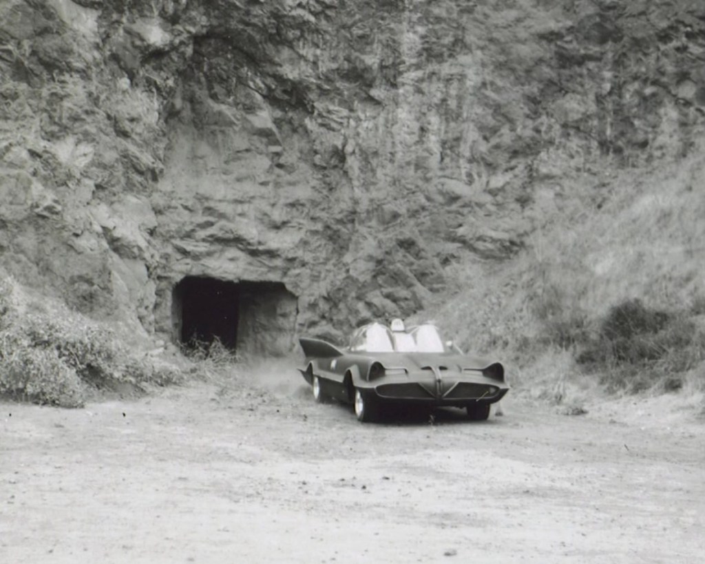 A black and white photograph of a sleek car positioned near a rocky cave entrance, suggesting a remote outdoor location.