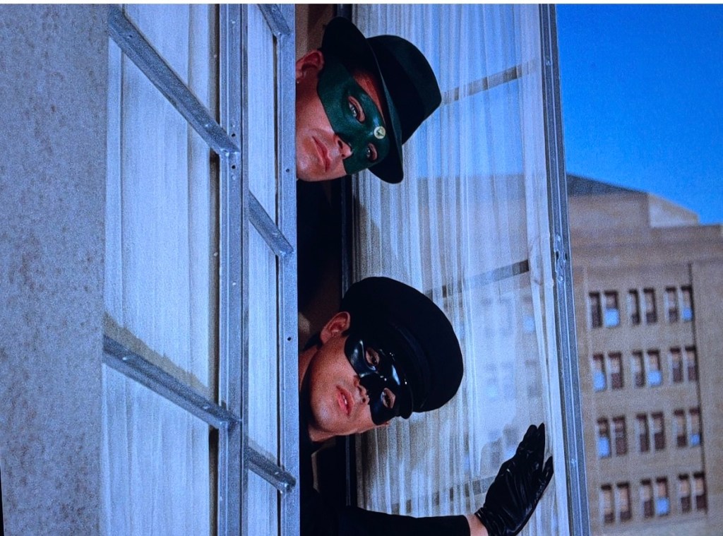Two masked characters peering out from a window, one wearing a black hat and the other a green hat, with a cityscape visible in the background.