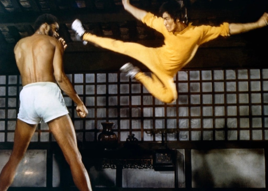 A scene depicting a martial arts duel, with one fighter in a yellow outfit performing a high kick above another fighter who is shirtless and positioned defensively.