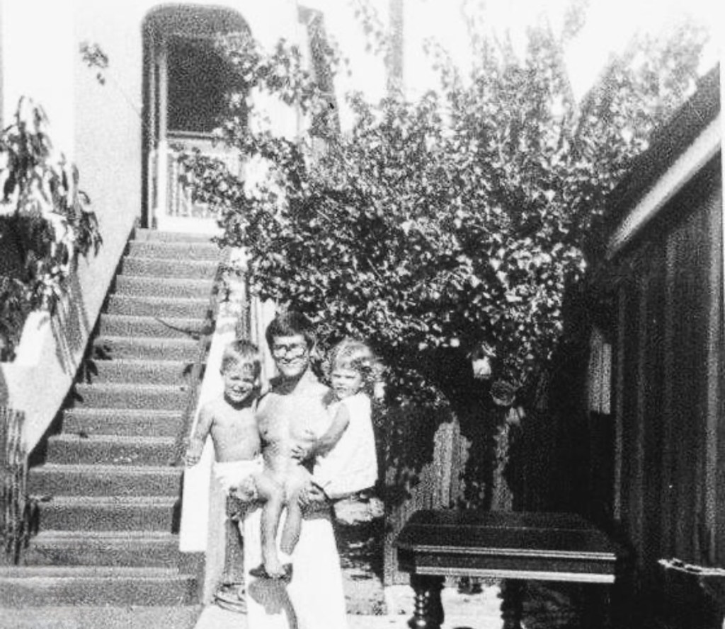 A black and white photo of a man holding two smiling children, standing outside a house with a staircase and a large bush in the background.