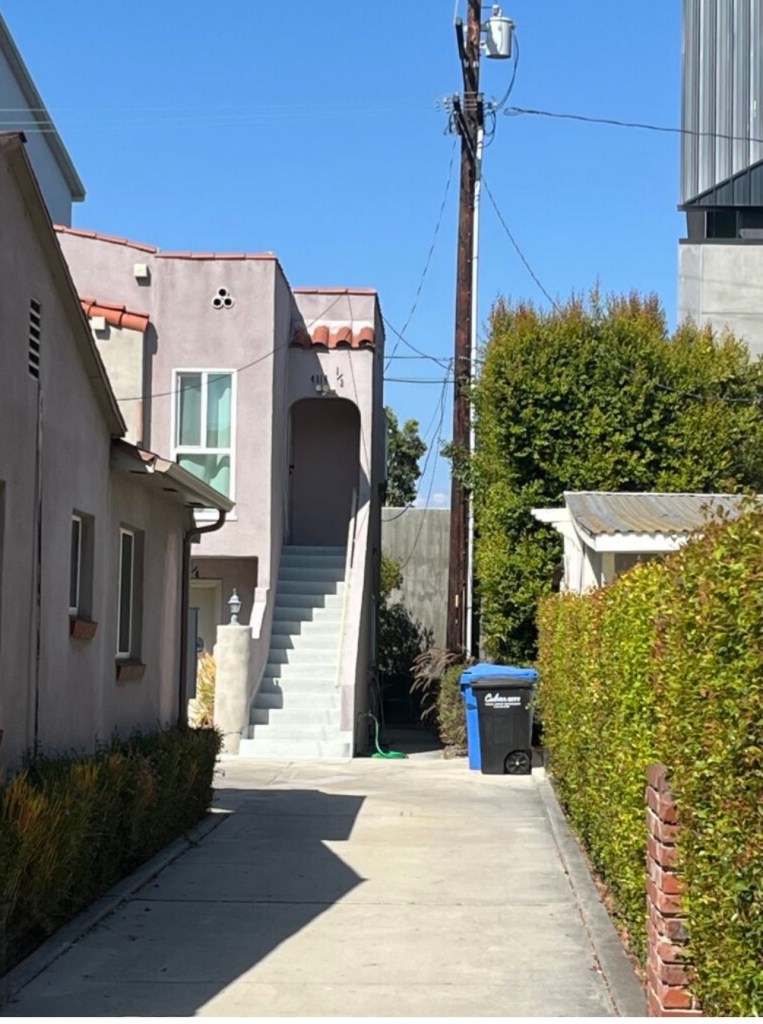 A narrow alleyway featuring two buildings with a staircase leading to an upper unit, flanked by hedges and a blue trash bin on the right side.