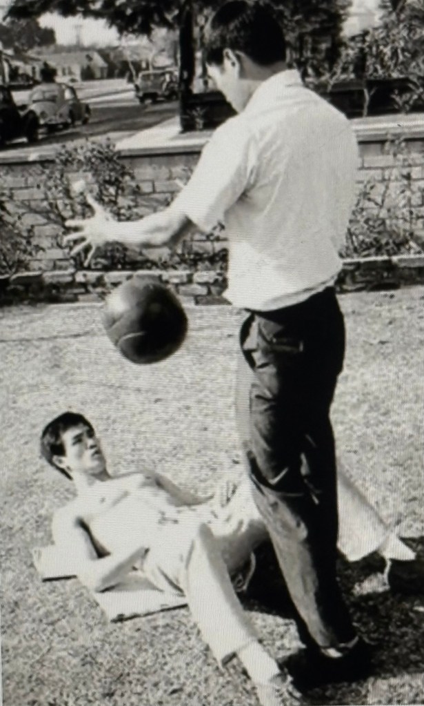 A man lying on a mat on the grass gazes up at another man, who is standing and preparing to drop a basketball towards him, in a black and white outdoor setting.