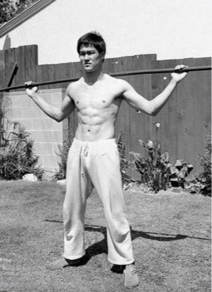 A black and white image of a muscular man standing in a garden, holding a stick with both hands. He is shirtless and wearing light-colored pants.