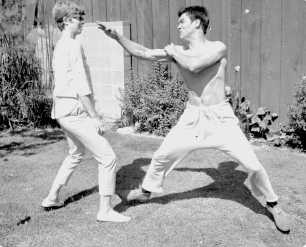 A black and white photo of a man in athletic wear demonstrating a martial arts move towards a woman, who is standing with a surprised expression. The background features a grassy area and wooden fence.