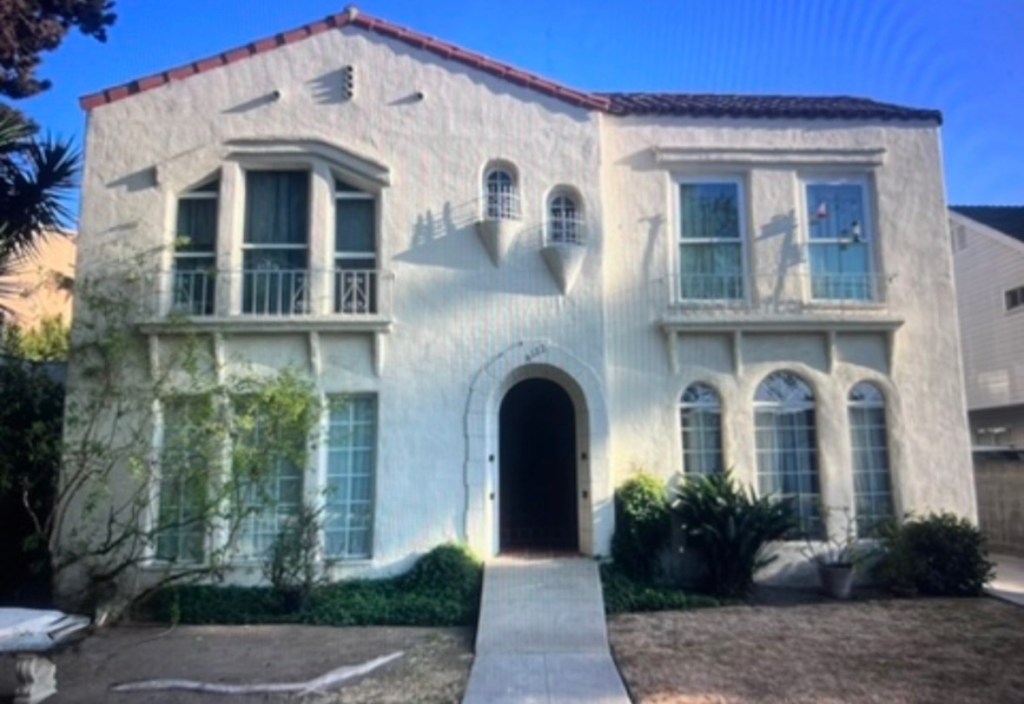A white, two-story residential building with a distinctive architectural style, featuring arched windows, a central entrance, and a tiled roof. The front yard includes greenery and a pathway leading to the entrance.