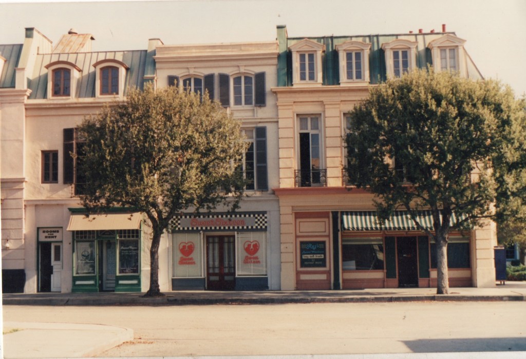 A street view of several buildings featuring a mix of architectural styles, with trees lining the sidewalk. The buildings include storefronts with signs, awnings, and various window designs.