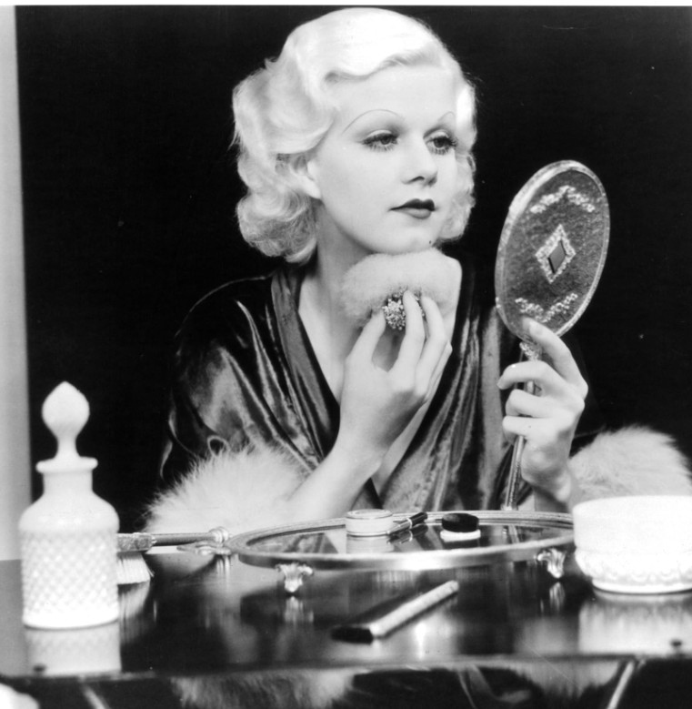 A glamorous woman in a silky robe using a powder puff while looking into a hand mirror, with vanity items visible on the table.