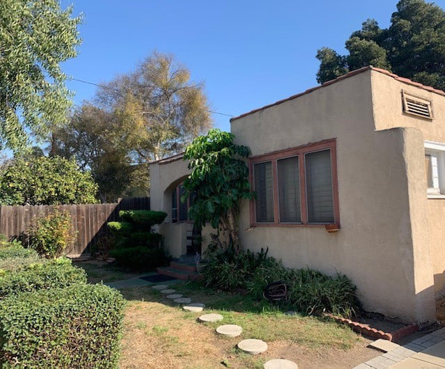 A small, beige house with a garden, featuring large windows and a pathway made of stone steps.