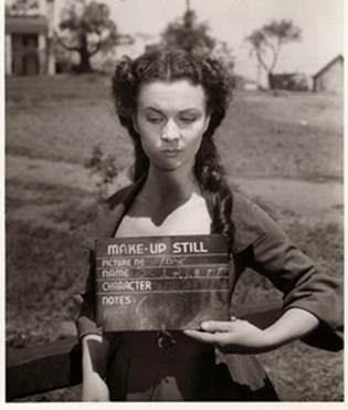 A woman holding a clapperboard labeled 'MAKE-UP STILL' with a serious expression, dressed in period clothing, outdoors with trees and a building in the background.