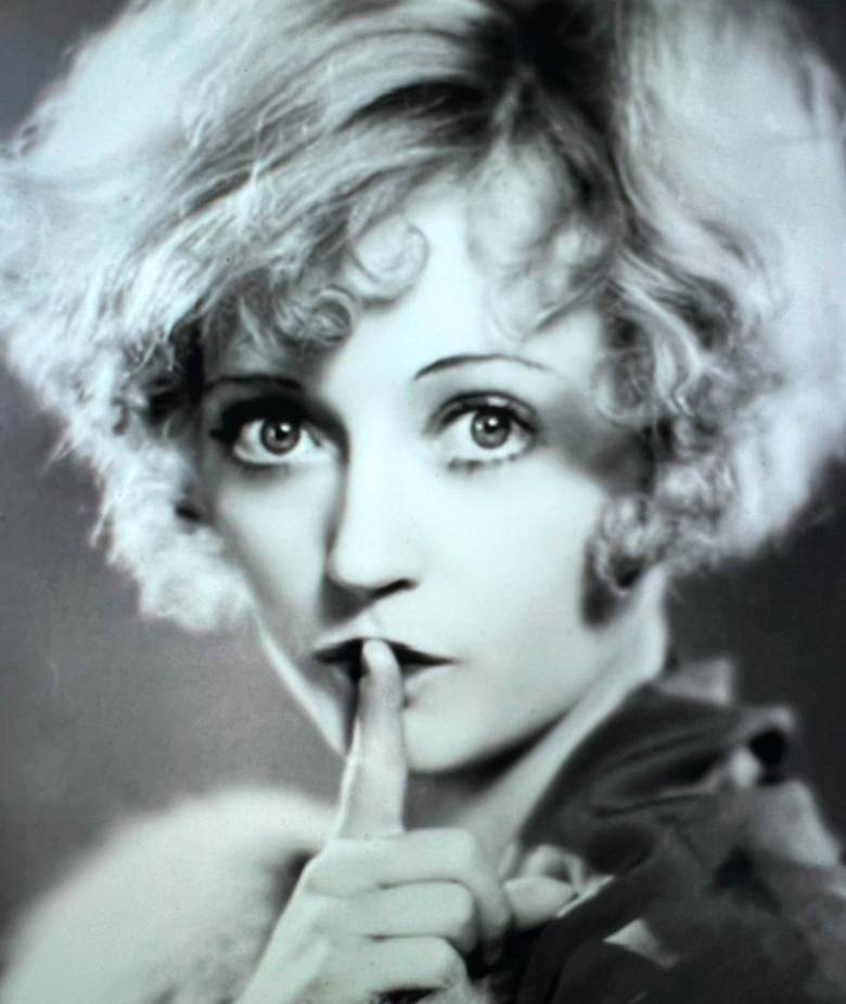 Black and white portrait of a woman with curly hair, making a 'shh' gesture with her finger to her lips.