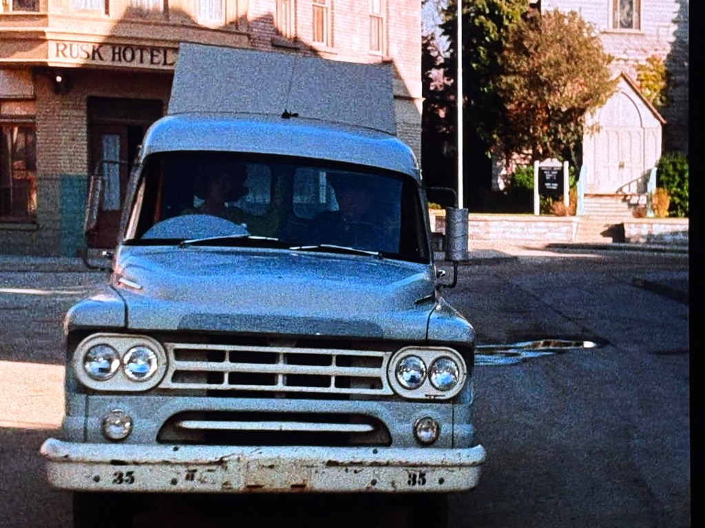 A vintage silver delivery truck with a boxy design parked in front of a historic hotel labeled 'Rusk Hotel'.