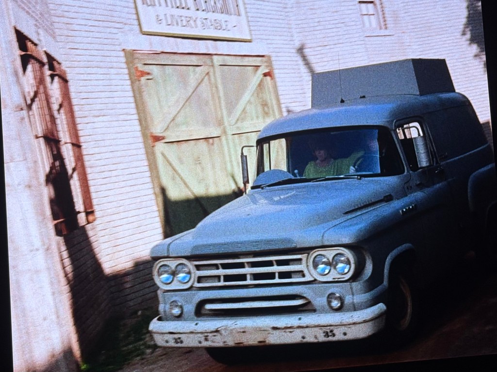 A vintage gray delivery truck driving past a rustic building with wooden doors.