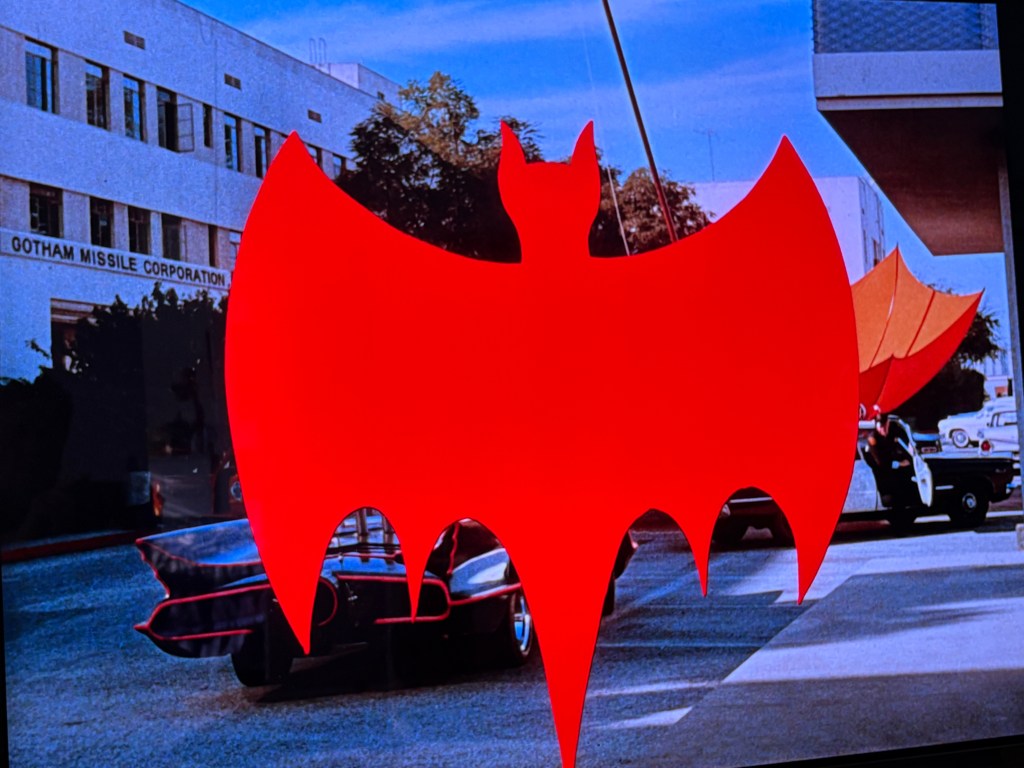 A bold red bat symbol in the foreground with buildings, vehicles, and a sign for Gotham Missile Corporation in the background.