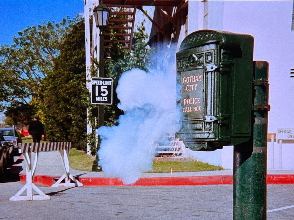 A green police call box labeled 'GOTHAM CITY POLICE CALL BOX' with smoke billowing from it, located on a street with a visible speed limit sign of 15 miles per hour and a police officer in the background.