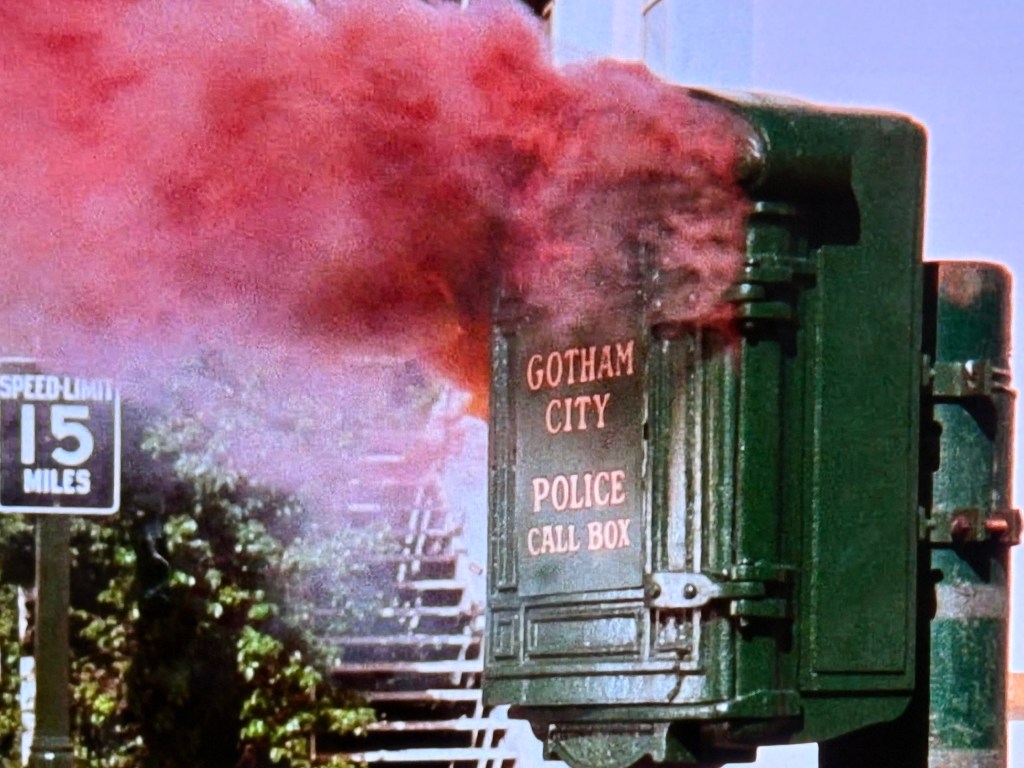 A Gotham City police call box emitting red smoke, with a speed limit sign reading 15 miles per hour in the background.