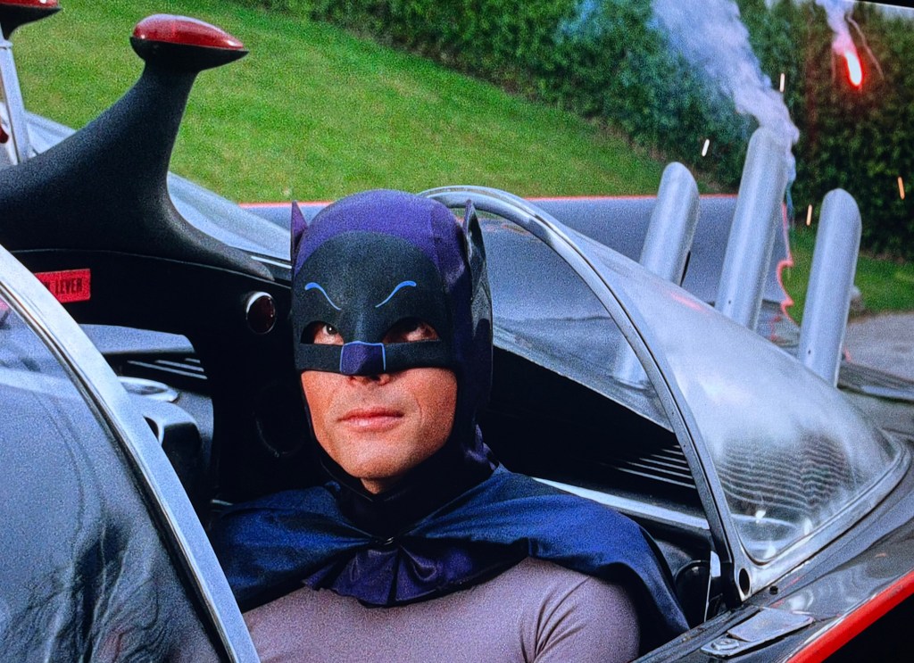 A character dressed as Batman, wearing a black mask and cape, sits in a classic Batmobile with visible rocket launchers in the background.
