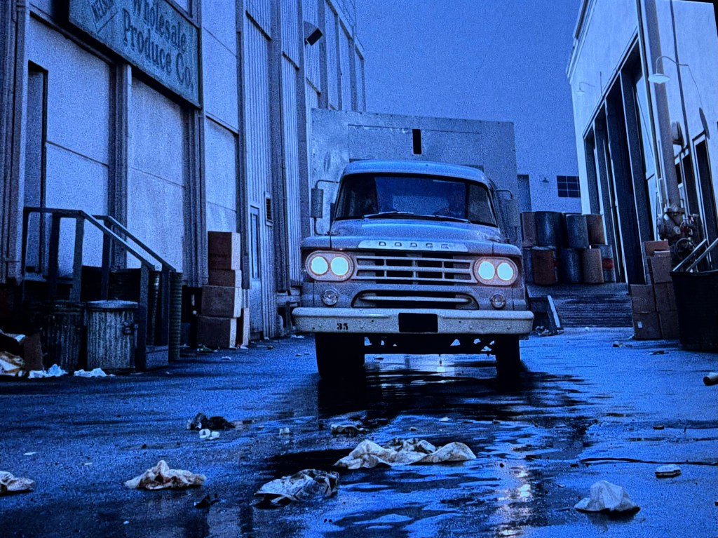 A vintage Dodge truck driving down a wet alley lined with garbage and boxes, illuminated by low light.