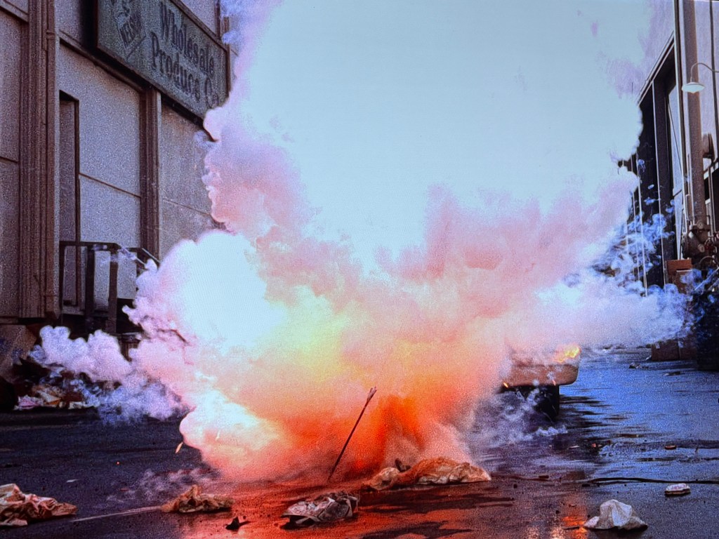 An explosion creating a large cloud of smoke and orange flames on a city street near a warehouse.