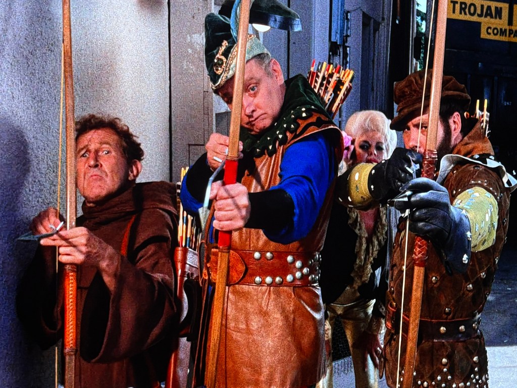A group of costumed characters, including archers and a woman, posing in a comedic manner. The scene features two men in medieval attire wielding bows, with another character partially visible in the background.