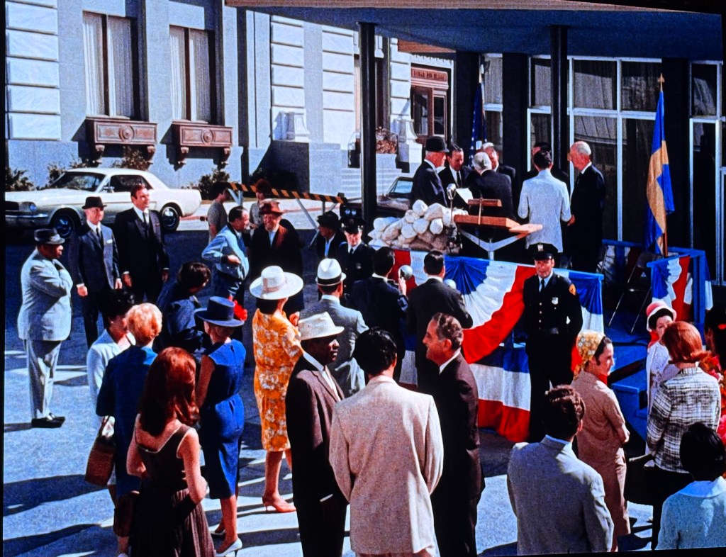 A crowd gathered outside a building, featuring people in formal attire and a police officer in uniform. There are flags and red, white, and blue decorations, suggesting a celebratory or official event.