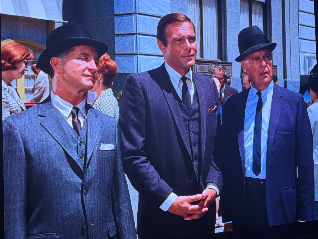 Three men in suits and hats standing together outdoors, with a crowd in the background.