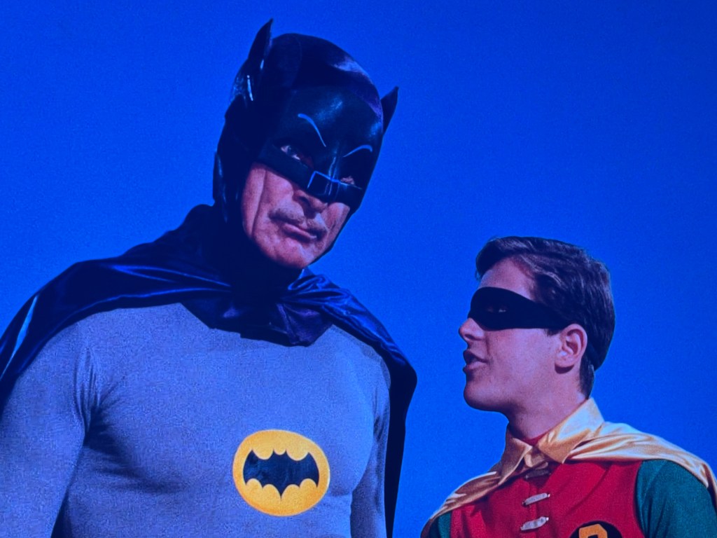 Batman and Robin stand together against a blue background, with Batman wearing his iconic suit and mask, and Robin in his colorful costume.