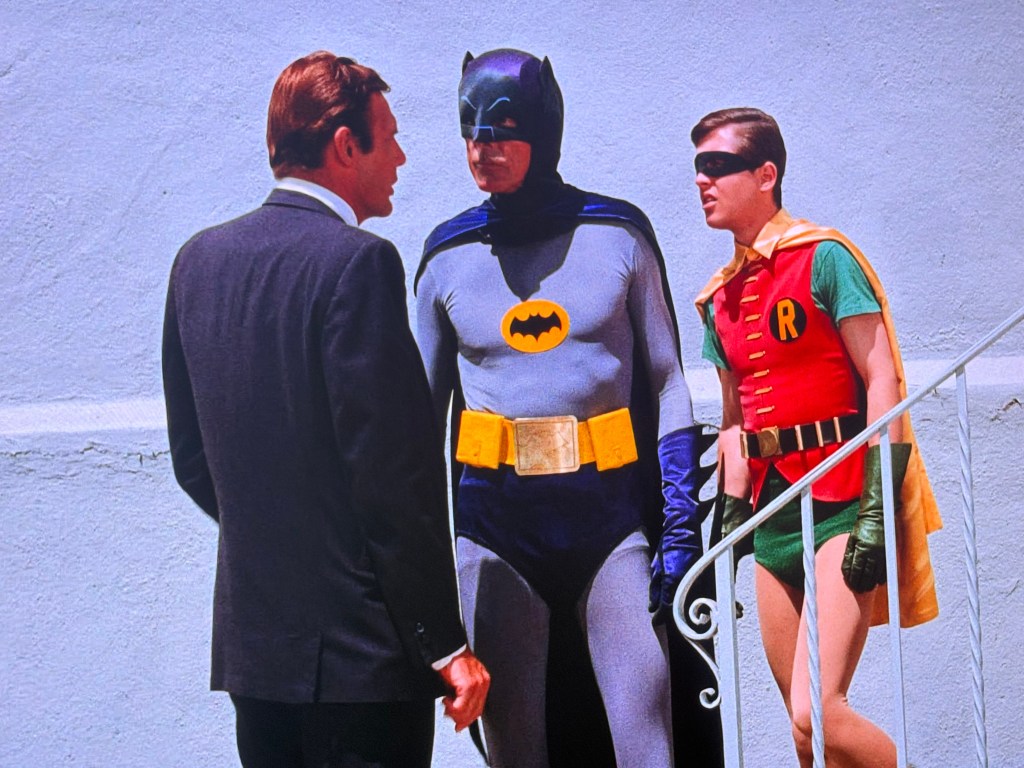 A scene featuring three characters: a man in a black suit speaking to Batman, who is in a grey suit with a yellow emblem, and Robin, dressed in a colorful costume with a cape.