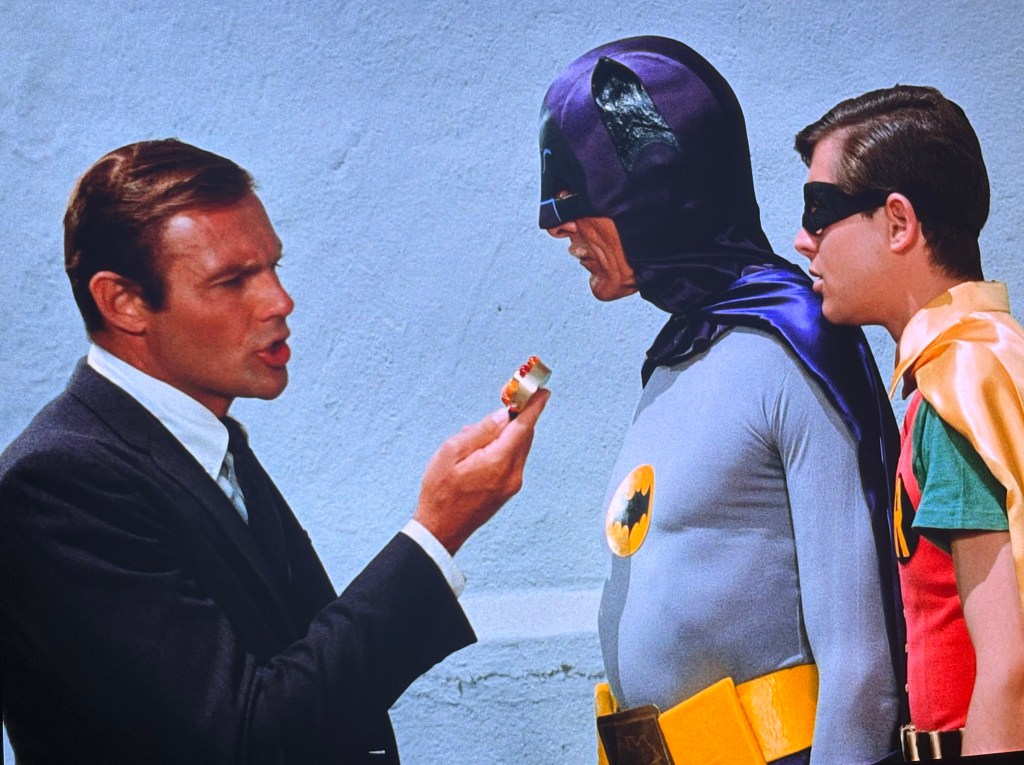 A scene featuring a man in a suit speaking animatedly to Batman and Robin, who are dressed in their iconic superhero costumes. The backdrop is a plain wall, and the man is holding a small object.