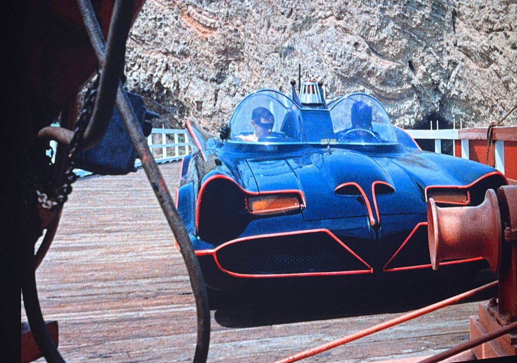 A classic black vehicle with red accents, resembling a futuristic car, parked on a wooden dock with rocky terrain in the background.