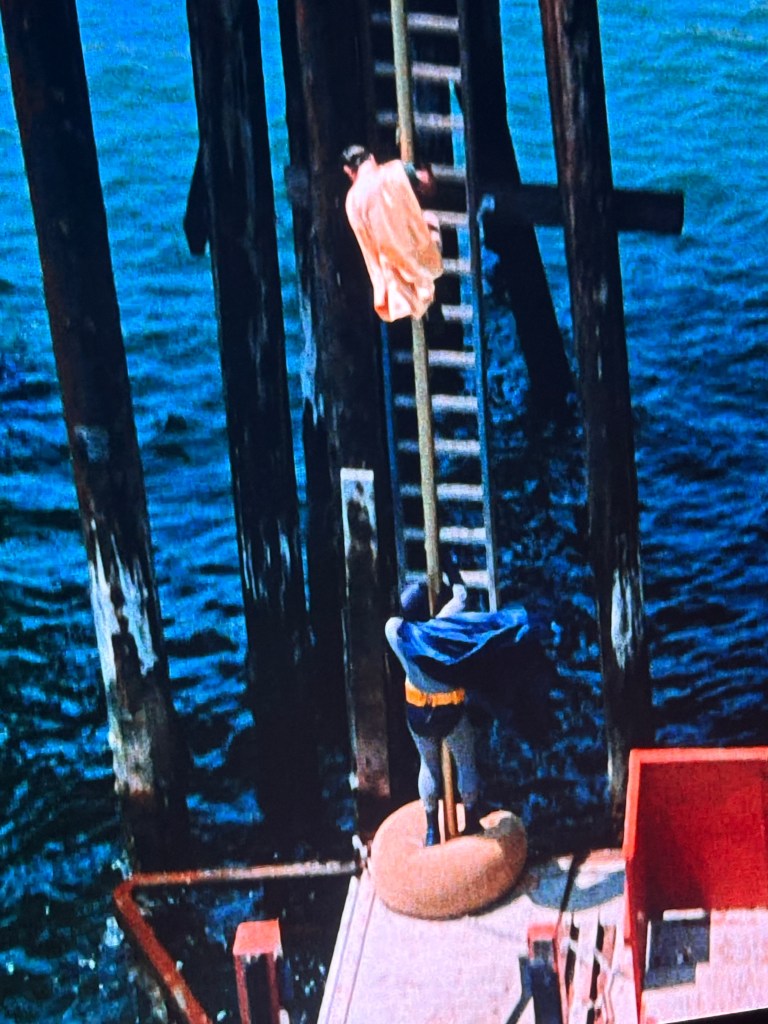 A superhero in a cape is positioned on a dock near a ladder leading up to a pier, while a figure in a light-colored outfit is seen climbing down the ladder.