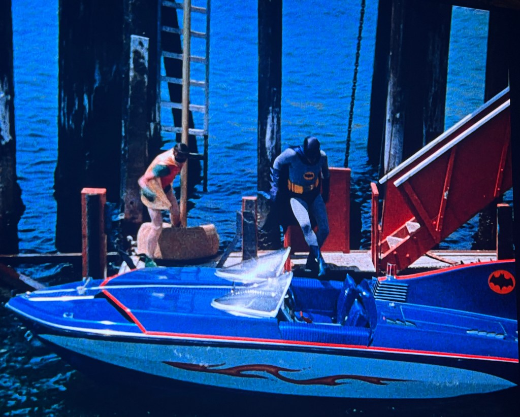 Batman and Robin at a dock near a blue speedboat with the Batman logo, with wooden pilings and a ladder in the background.