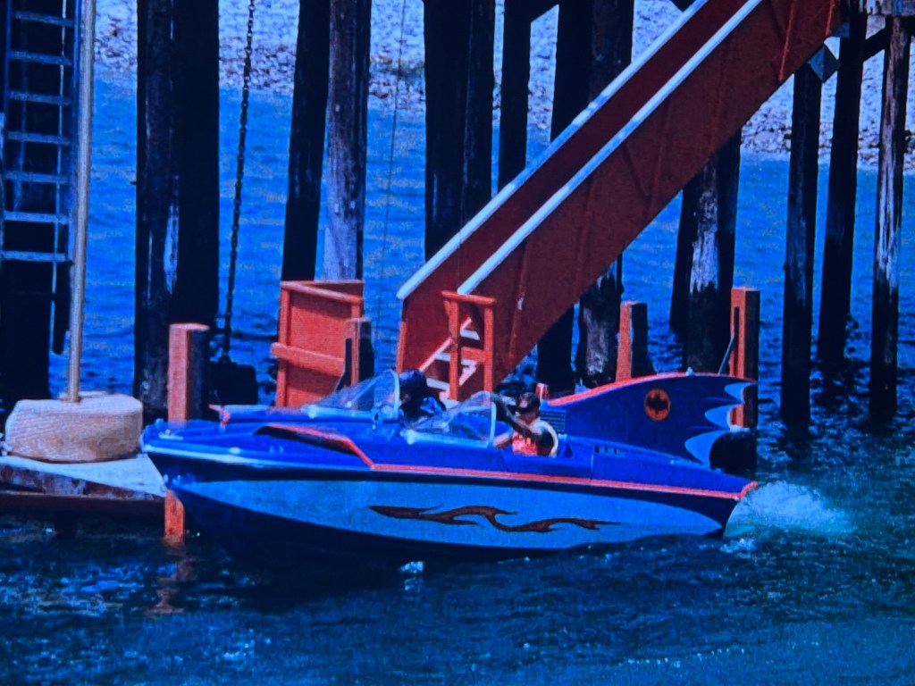 A colorful speedboat, featuring a design with flames and a bat logo, approaches a dock with wooden pillars and a red structure in the background.